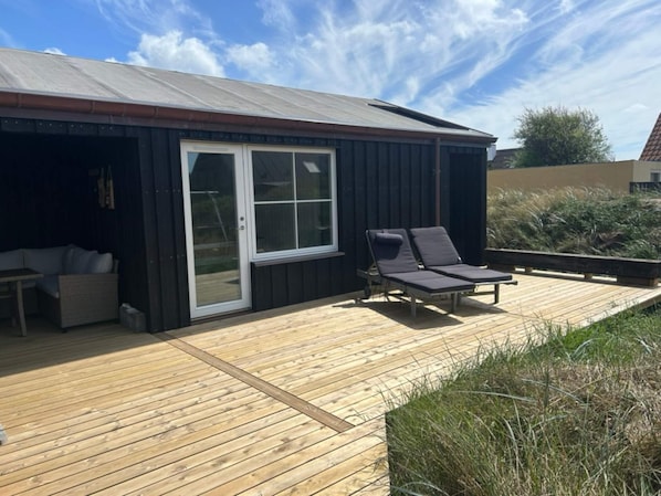 A wooden deck with two lounge chairs, a small table, and a sliding glass door leading to a covered area.