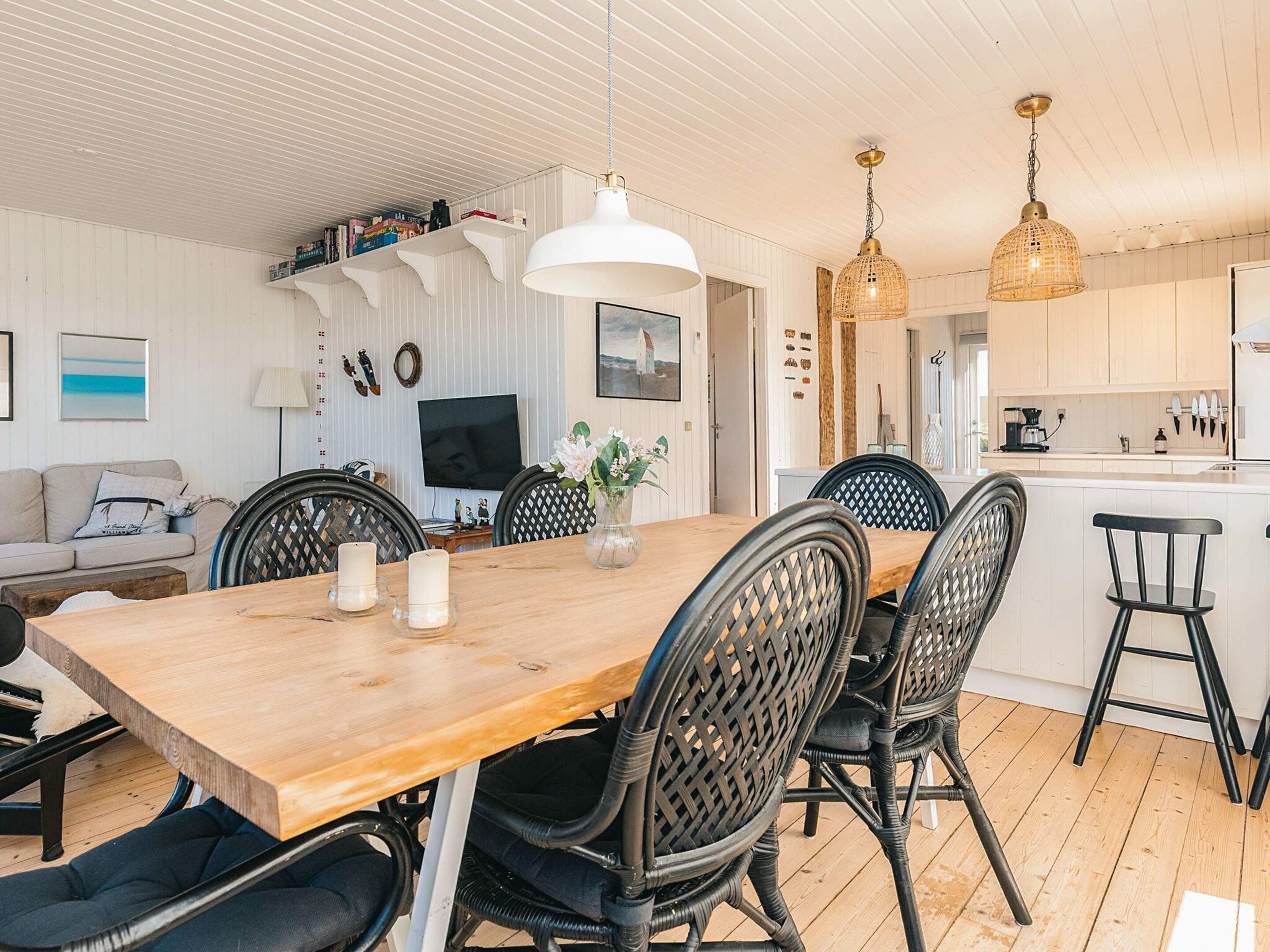 A modern living room with a wooden dining table, black chairs, a TV, and a kitchen area with white cabinets.