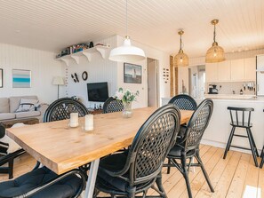 A modern living room with a wooden dining table, black chairs, a TV, and a kitchen area with white cabinets. - 4 Star Holiday Home in Skagen (Skagen)