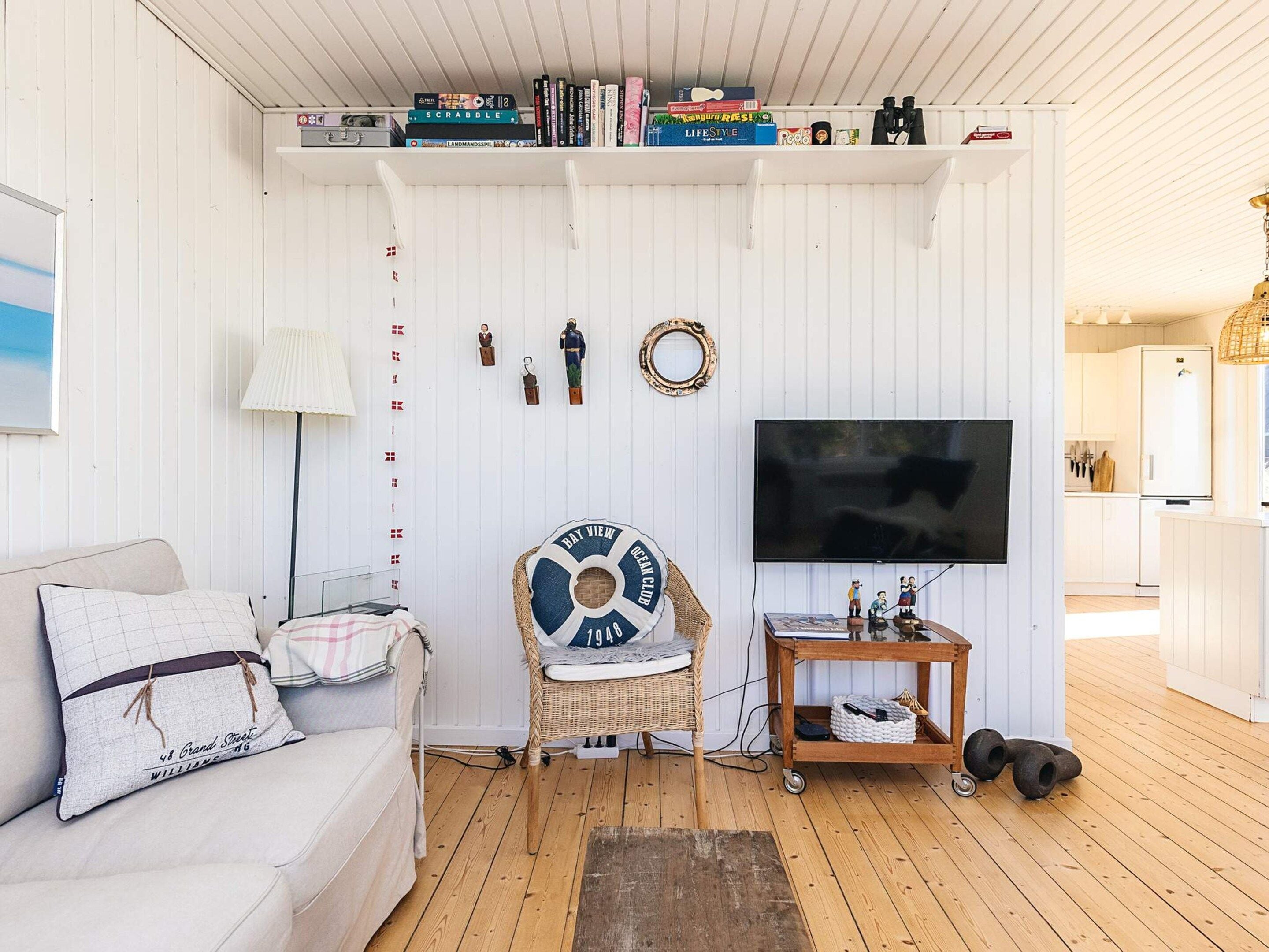 Een woonkamer met een witte muur, een televisie, een bank met patroon kussens, een rieten stoel en een boekenkast met diverse objecten.