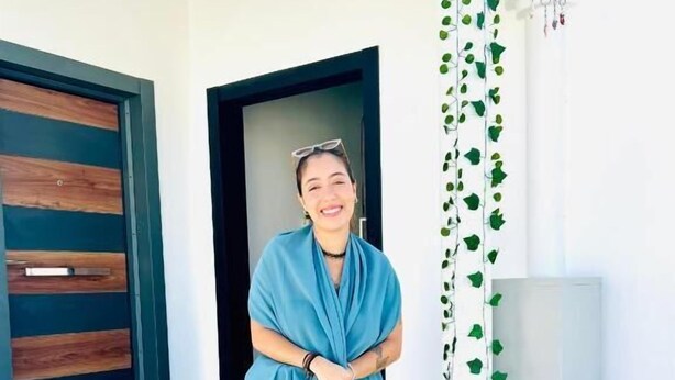Una persona parada en una escalera frente a la entrada de un edificio moderno, que cuenta con una puerta de madera y un pilar blanco con follaje verde.