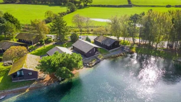 A lakeside property with several buildings, including a main house and smaller structures, surrounded by lush greenery and a calm lake. - Swanmere lodge, lakeside view cabin at Pinelake Carnforth (Carnforth)