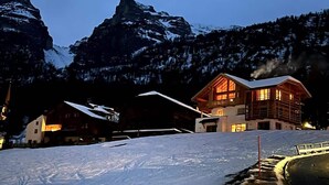 Een bergchalet met een grote houten constructie, een kleiner bijgebouw en een besneeerd landschap met bergen op de achtergrond.