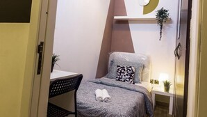 Quarto de hotel pequeno, com uma cama, uma mesinha e uma cadeira. Há uma prateleira na parede com um relógio e uma planta.