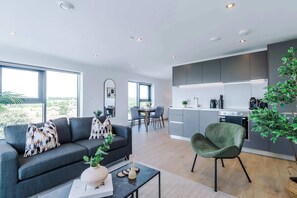 A modern living room with a grey sofa, a green armchair, and a dining area with a table and chairs. A kitchen with grey cabinets and a stove is visible in the background. - Premium City Penthouse Ideal For Business (Leeds)
