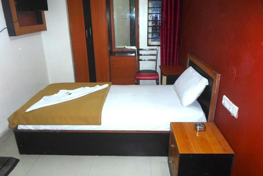 A single bed with a brown bedspread and white sheets, a wooden nightstand with a lamp, a red chair, and a small table with a tissue box.
