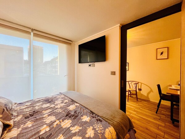 A hotel room with a bed, a TV on the wall, a window, and a door leading to a dining area. - Buho Urban Curic A C Washer Dryer Wifi (Santiago)