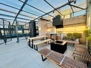 An outdoor patio with wooden benches and tables, a grill, and a TV mounted on a brick wall. - Buho Urban Curic A C Washer Dryer Wifi (Santiago)