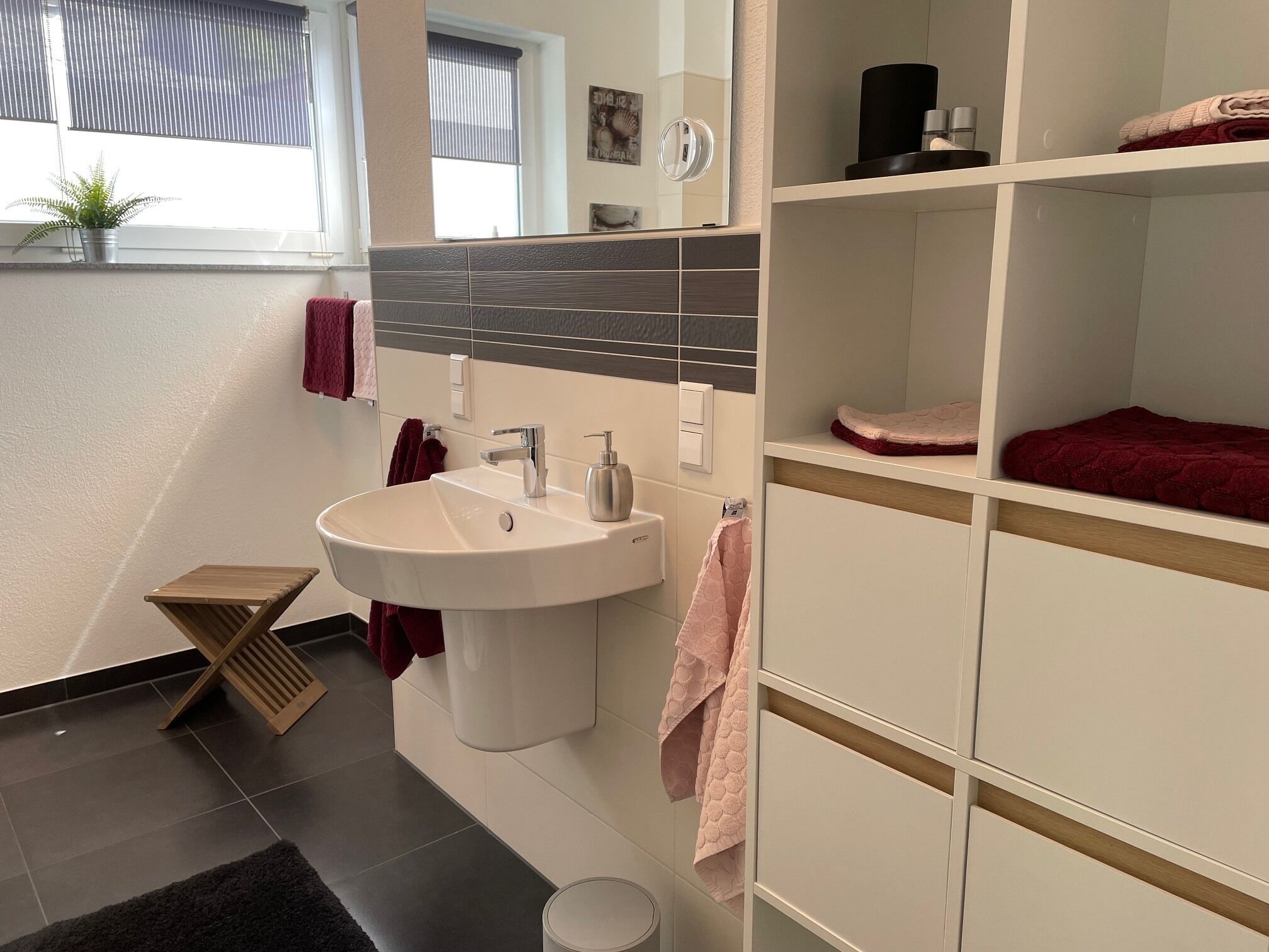 A modern bathroom with a large sink, a mirror, and a towel rack. There is a small wooden stool and a black rug on the floor.