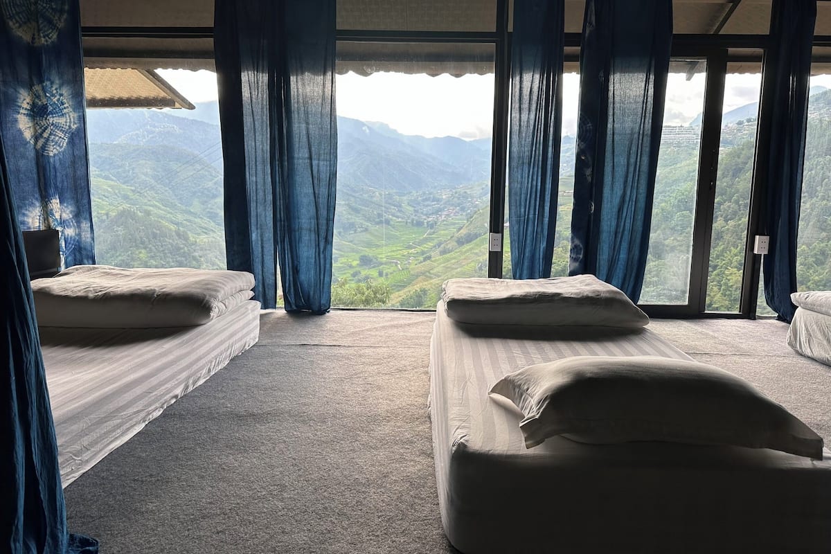 Une chambre avec deux lits, un sol recouvert de moquette et une vue sur des collines verdoyantes, visible à travers de grandes fenêtres.