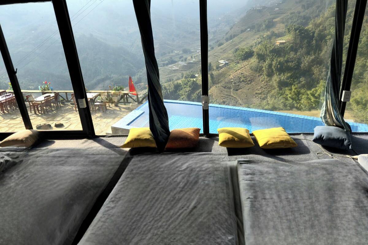 Une pièce dotée d’une grande fenêtre donnant sur un paysage montagneux, une piscine, ainsi qu’une table avec des chaises et une plante.