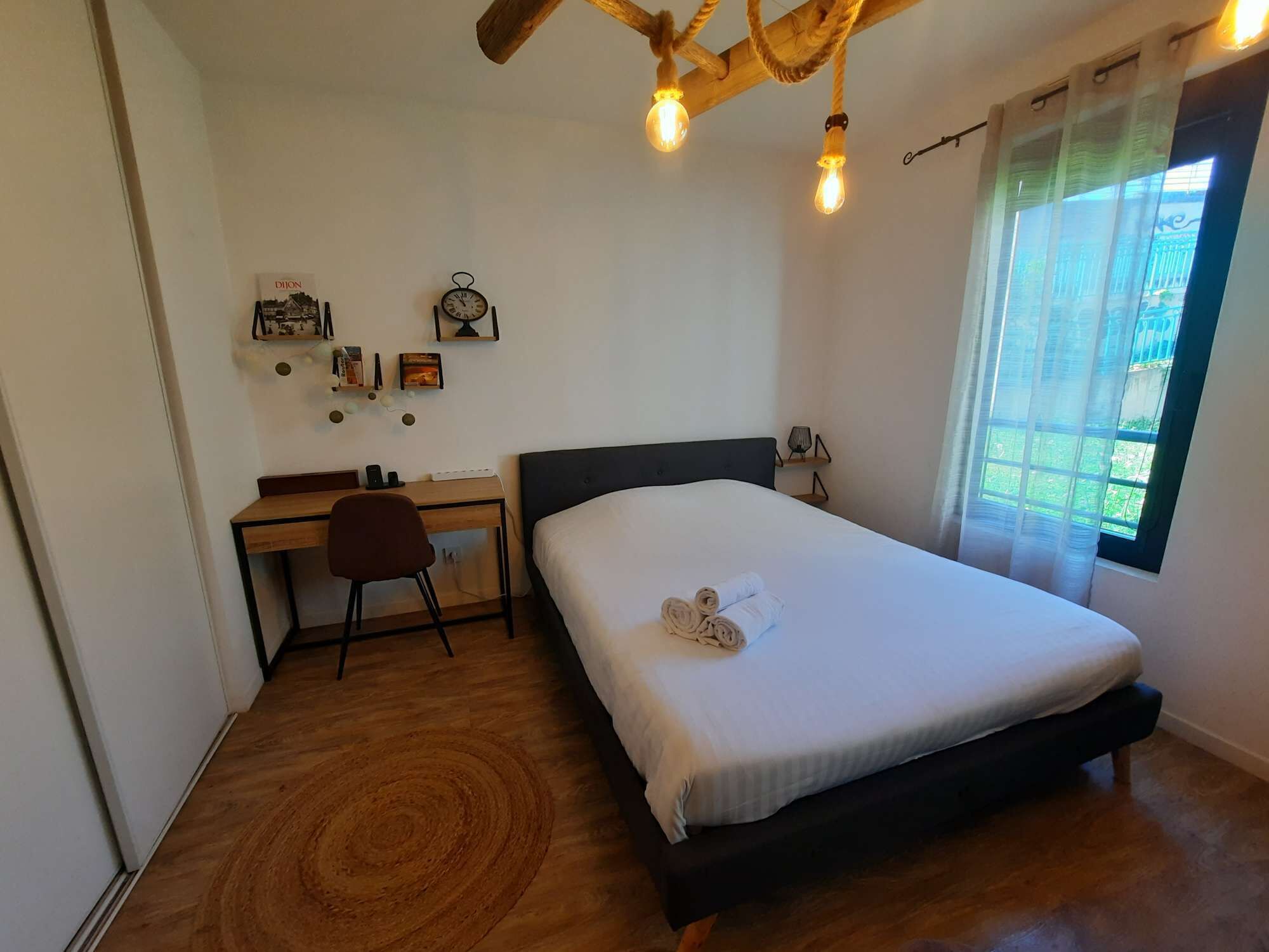 A bedroom with a bed, a desk, and a chair. There are wall decorations and a window with curtains.