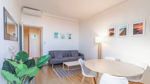 Una sala moderna con un sofá gris, una mesa redonda y una lámpara de pie. Hay cuadros enmarcados en la pared y una planta verde grande en la esquina.