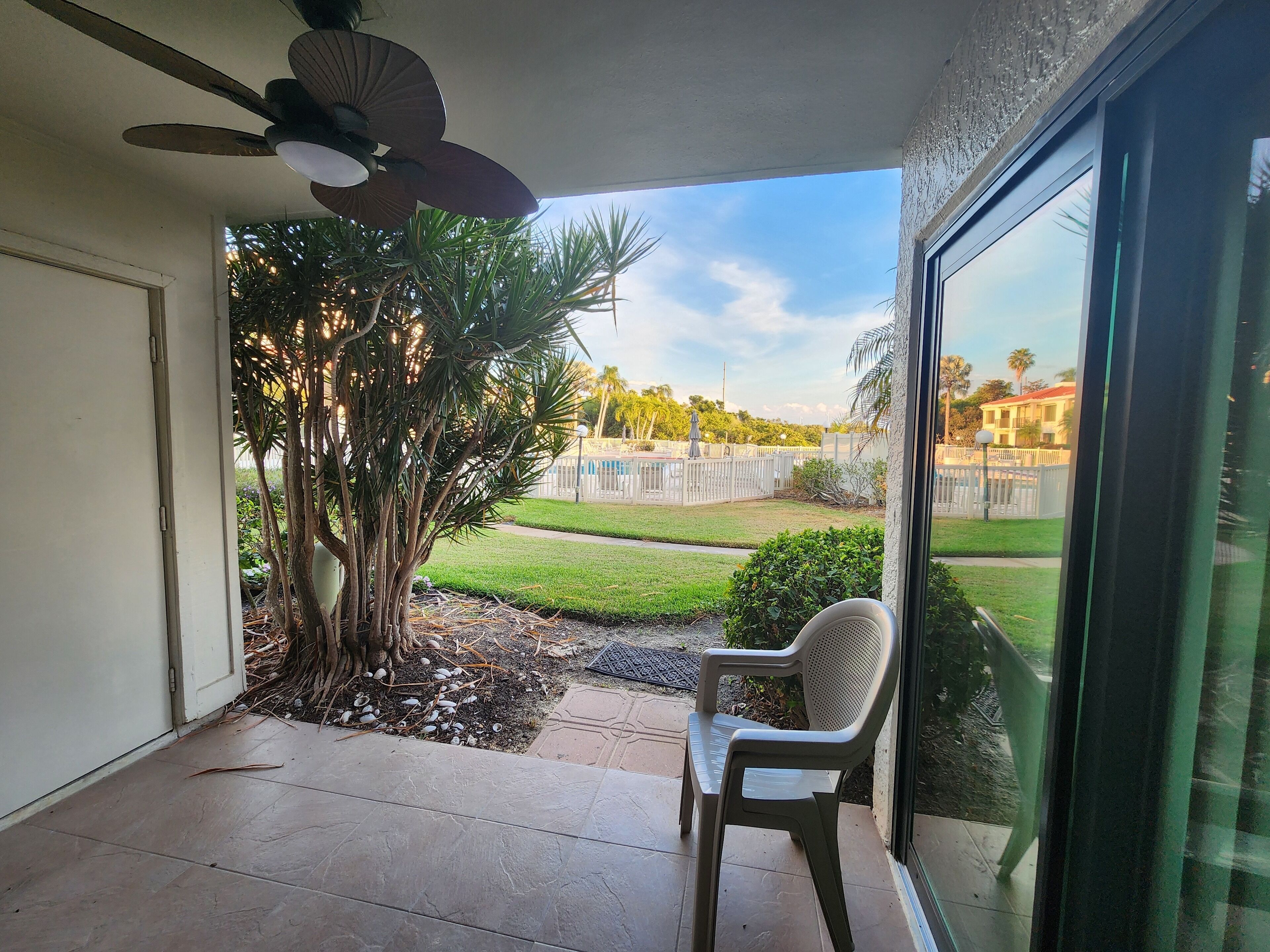 A patio with a ceiling fan, a chair, and a view of a well-maintained lawn and distant buildings.