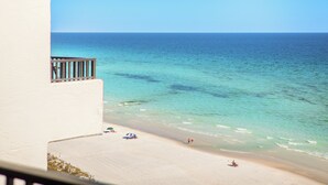 En strand med utsikt över klart vatten, en sandstrand och en vit byggnad med en balkong.