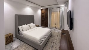 A modern bedroom with a large bed, a wooden wardrobe, a TV mounted on the wall, and a window with sheer curtains. - East Legon Hills, Accra, Nanakrom Gated Community (Accra)