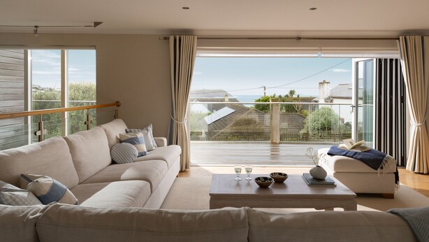 A modern living room with a large window, a sofa, a coffee table, and a balcony with a view of the sea. - Springtide - New Polzeath - stylish sea view property sleeping 8-9 guests (New Polzeath)
