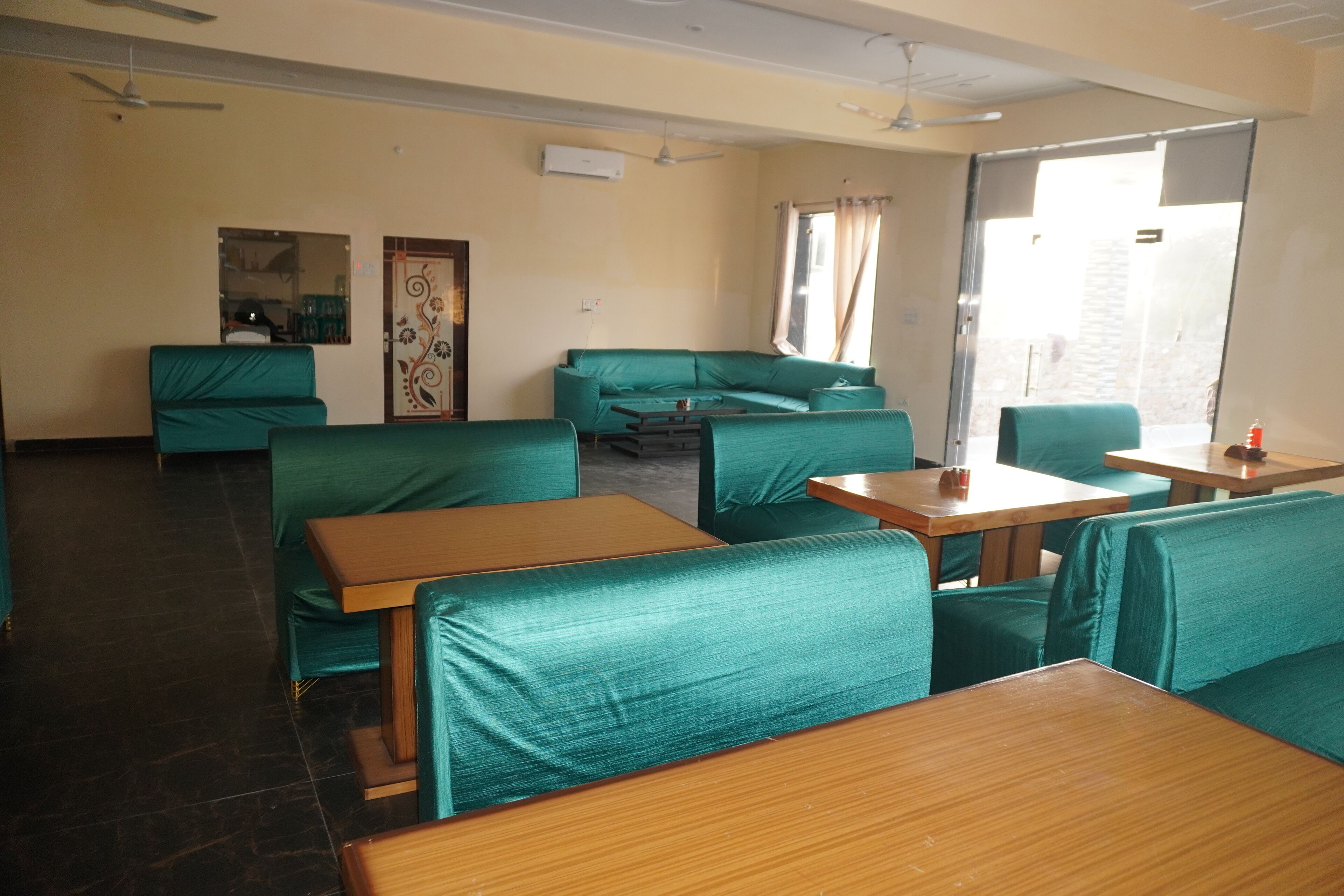 A room with green upholstered seating, wooden tables, a ceiling fan, and a window with curtains.