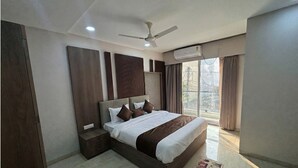 A modern bedroom with a large bed, a nightstand, a ceiling fan, and a view of the outdoors through a large window. - Hotel Shaurya Palace (Indore)