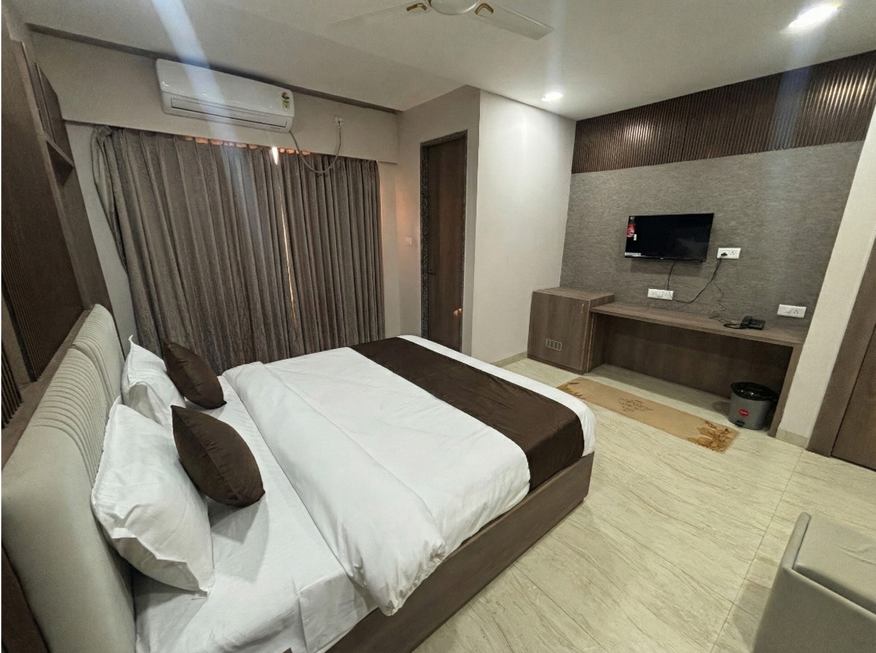 A modern hotel room with a large bed, a flat-screen TV mounted on a wall, a small cabinet, and an air conditioning unit.