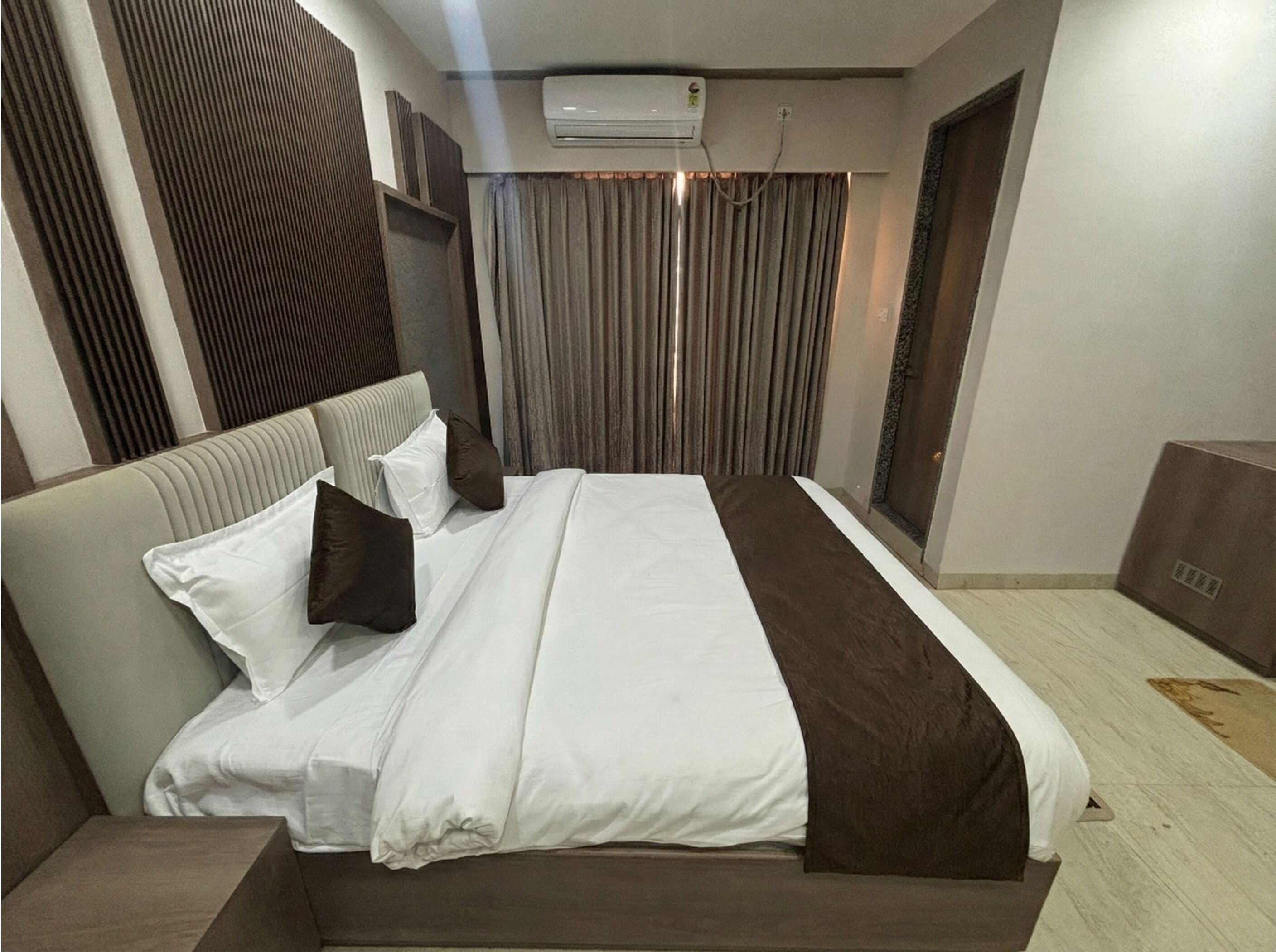 Une chambre d’hôtel comprenant un lit, une tête de lit, une fenêtre avec des rideaux, un appareil de climatisation et un plancher en bois.