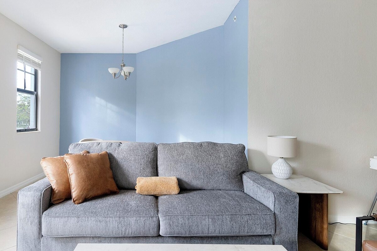 A living room with a grey sofa, two brown cushions, a white side table with a lamp, and a ceiling light fixture.