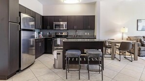 A modern kitchen with dark cabinets, stainless steel appliances, a central island with bar stools, and a dining area with a table and chairs. - Landing Furnished Apartments at Miramar Town Center (Miramar)