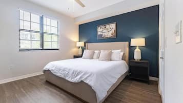 Un dormitorio con una cama grande, dos mesitas de noche con lámparas, un ventilador de techo, una ventana con persianas y una obra de arte colgada en la pared.