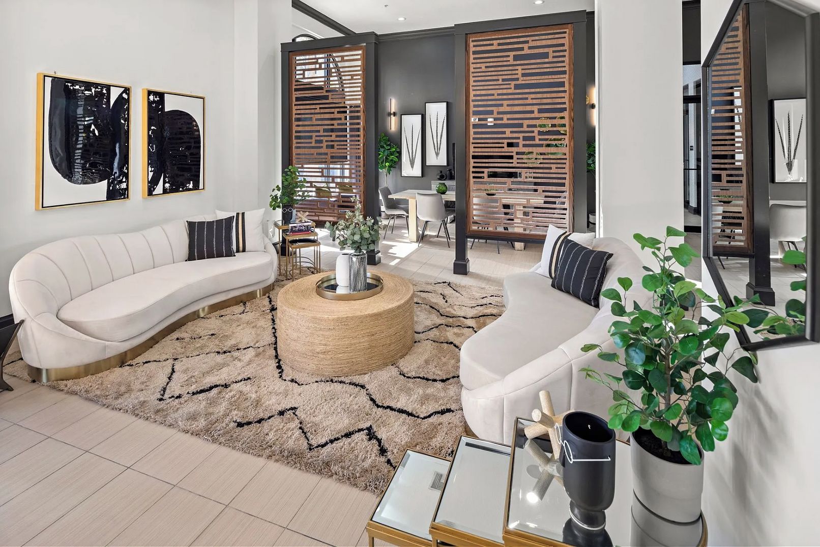 A modern living room with a curved white sofa, a round coffee table, and a large mirror. There are two wooden partitions with plants, and a dining area is visible in the background.