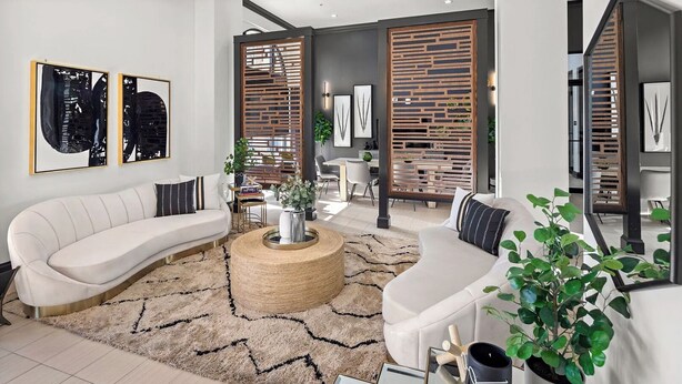 A modern living room with a curved white sofa, a round coffee table, and a large mirror. There are two wooden partitions with plants, and a dining area is visible in the background. - Landing Furnished Apartments at Miramar Town Center (Miramar)
