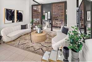 A modern living room with a curved white sofa, a round coffee table, and a large mirror. There are two wooden partitions with plants, and a dining area is visible in the background. - Landing Furnished Apartments at Miramar Town Center (Miramar)