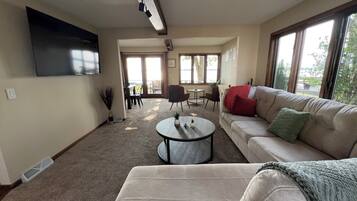 A living room with a sofa, a coffee table, and a TV mounted on the wall. There is a dining area with chairs and a table, and large windows providing natural light.