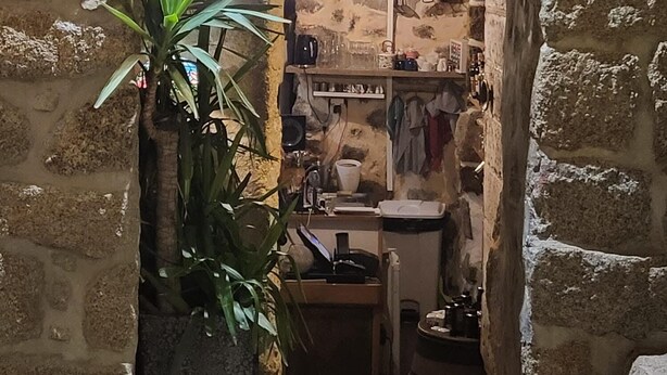 Um arco de pedra com uma placa indicando "La Vieille Ville". No interior, há prateleiras com diversos objetos e um lustre pendurado.