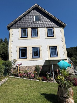 A two-story house with a gabled roof, multiple windows, and a well-maintained garden with colorful flowers and a large fish sculpture. - Landhaus Sommerberg - Fresh Forest Air, Nature & Leisure Apartment II, Thuringian Forest (Schleusingen)