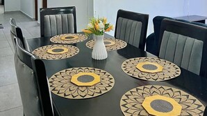 Un comedor con una mesa negra, seis sillas y un florero con flores. Hay luminarias fijadas a la pared y un televisor en la pared.