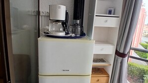 Ett litet rum med ett kylskåp från Waltham, en kaffemaskin, en liten hylla, ett fönster med gardiner och utsikt över grönområden utanför.