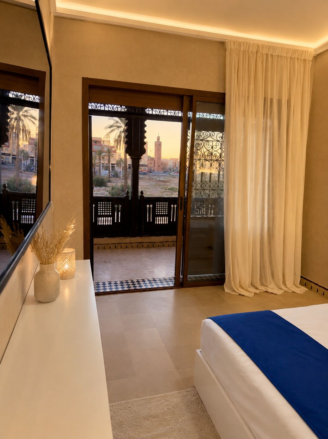 A hotel room with a bed, a desk, a vase with dried plants, a lamp, and a view of the cityscape through a glass door.
