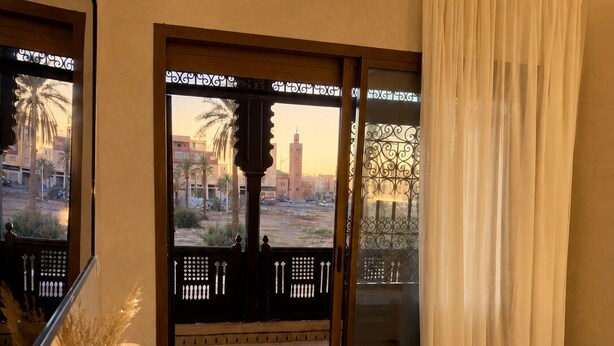 A hotel room with a bed, a desk, a vase with dried plants, a lamp, and a view of the cityscape through a glass door. - Cosy villa targa garden 5G IPTV (Marrakech)