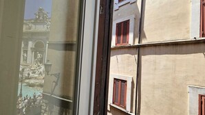 Blick durch ein Fenster auf ein Gebäude mit steinverkleideter Fassade und roten Fensterläden, mit Blick auf einen öffentlichen Platz mit Springbrunnen und Statuen.