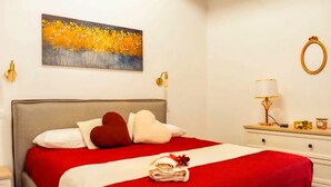 Ein ordentlich bezogenes Bett mit einer rot-weißen Tagesdecke, einem herzförmigen Kopfkissen und einem gerollten Handtuch mit roter Schleife. Über dem Bett hängt ein Gemälde von gelben Blumen.