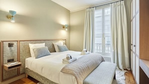 A bedroom with a large bed, bedside tables, a bench, a window with curtains, and wall-mounted lights. - Luxurious Apartment AC Saint Germain des Pres (Paris)
