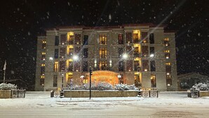 A large, multi-story building with illuminated windows and a central entrance, surrounded by snow-covered ground and a few snowbanks. - ILK INN HOTEL (Qusar)