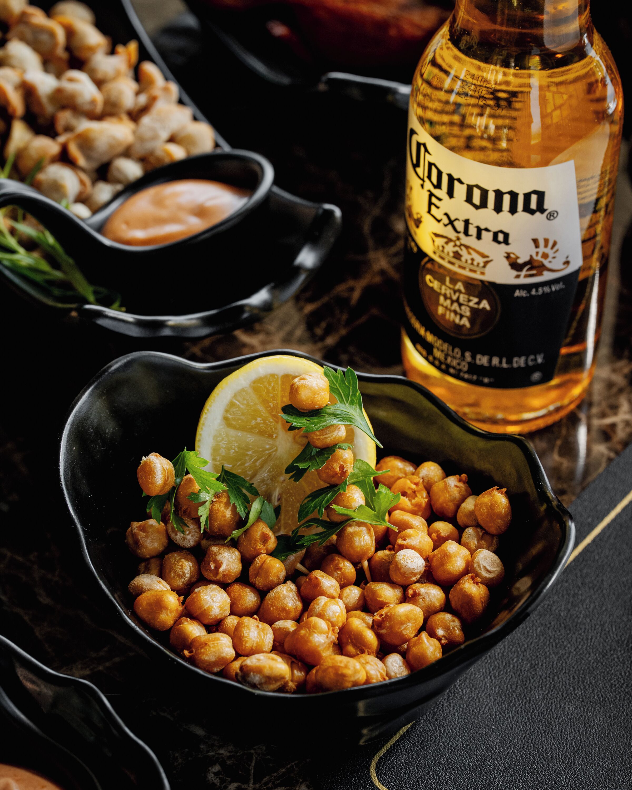 Un tazón de garbanzos adornado con perejil y limón, con una botella de cerveza Corona Extra al fondo.