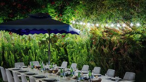 An outdoor dining area with tables set for six, surrounded by lush greenery and a blue umbrella. - ILK INN HOTEL (Qusar)