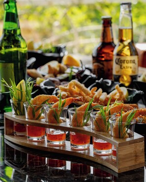 Un plateau de hors-d’œuvre aux crevettes et au romarin, accompagné de petits verres contenant une boisson rouge, et des bouteilles de bière en arrière-plan.