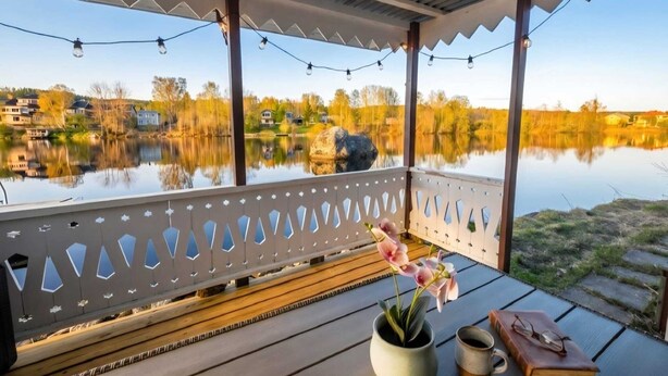 A covered deck with a view of a lake and houses, a potted plant, and a book and glasses on a table. - Riverside Villa with a Private Jetty and Scenic Views (Torpshammar)