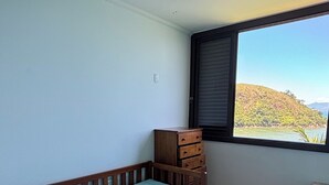 Et soveværelse med en enkelt seng, en trækommode, en ventilator i loftet og et vindue med udsigt over et bakket landskab.