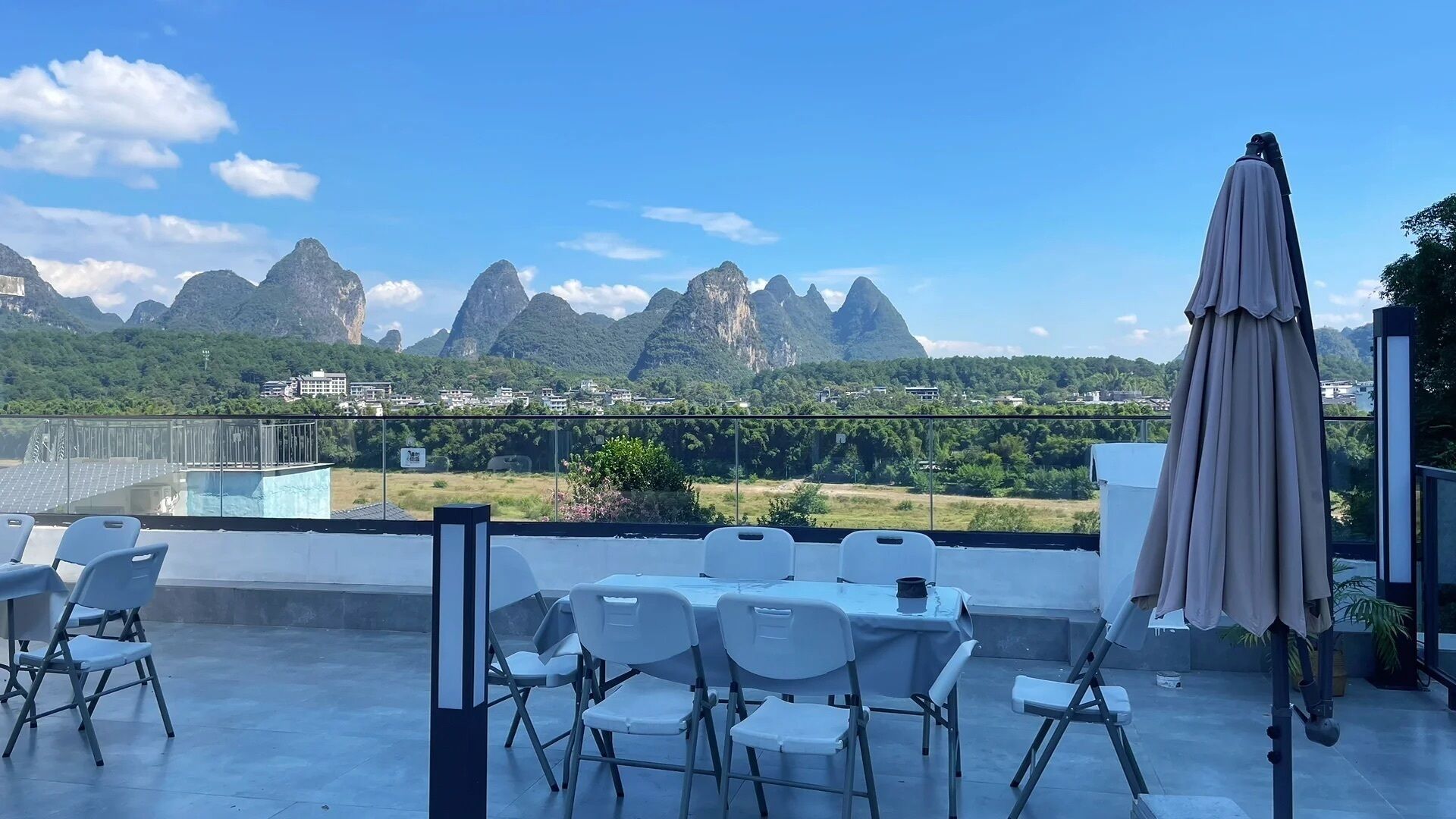 A terrace with chairs, a table, and an umbrella, overlooking a mountainous landscape with a pool and buildings in the background.