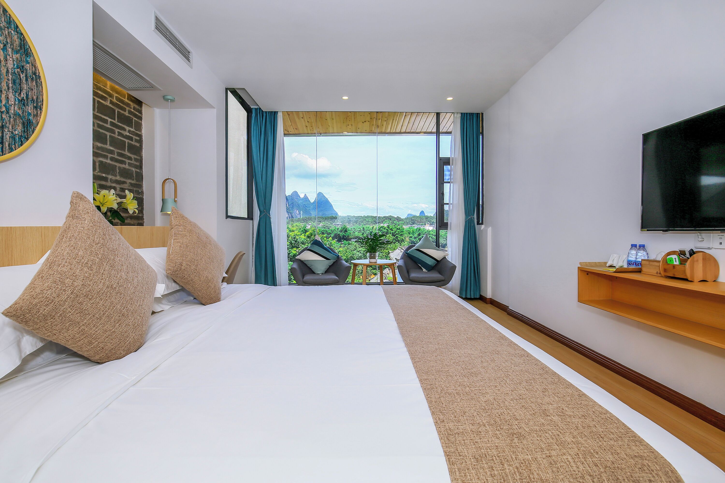 A hotel room with a large bed, a TV, a small table with a plant, and a view of mountains through the window.