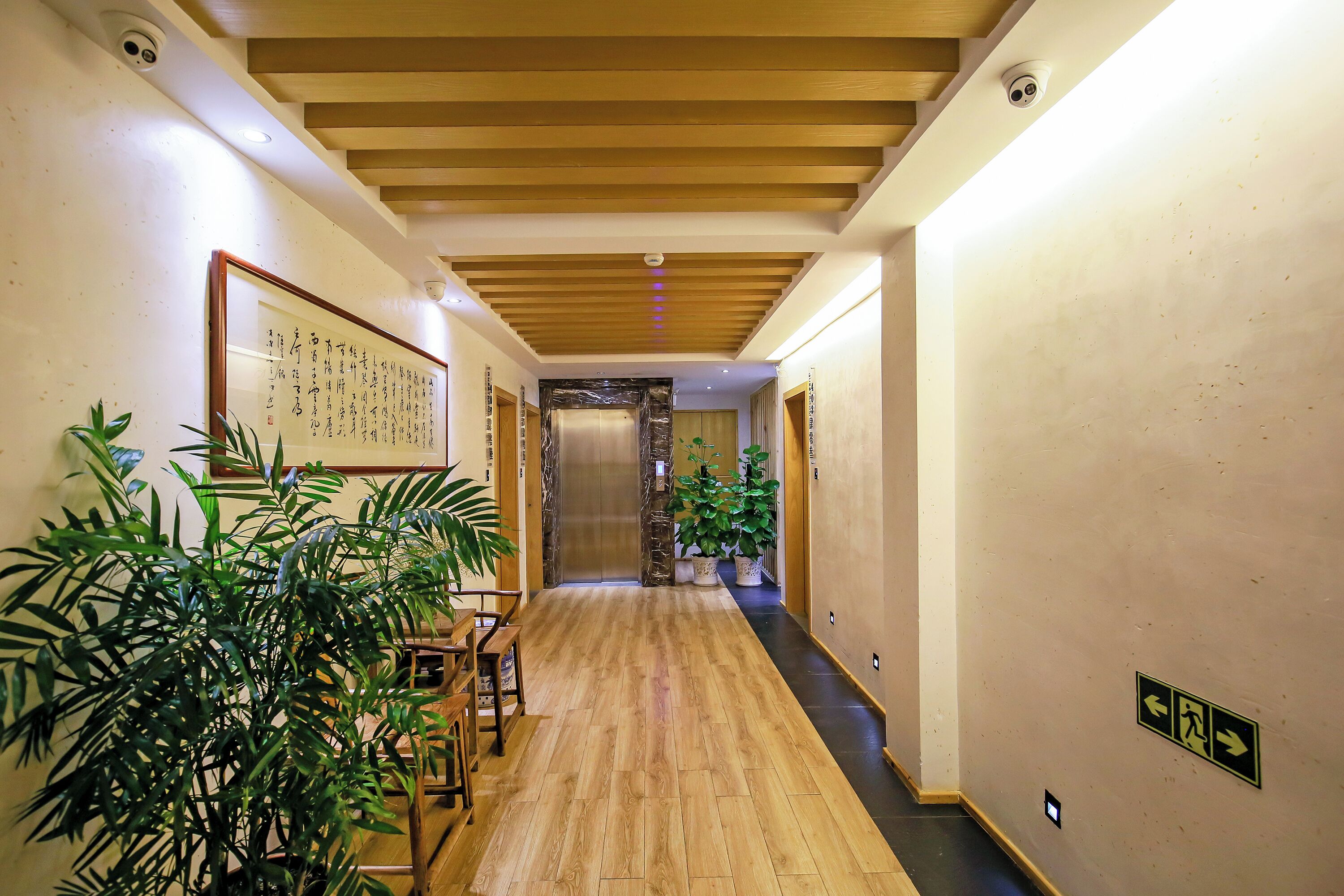 A hallway with wooden flooring, potted plants, a framed calligraphy piece, and an elevator.